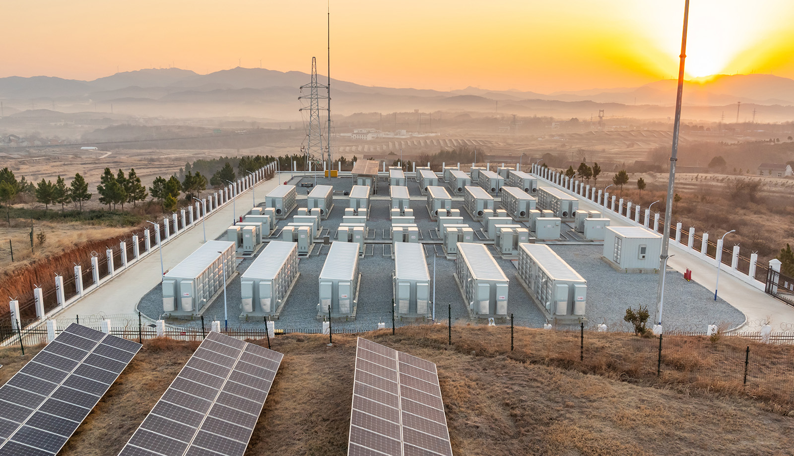 Große Batteriespeicheranlage mit Solarpanels im Vordergrund bei Sonnenaufgang, eingebettet in eine hügelige Landschaft.
