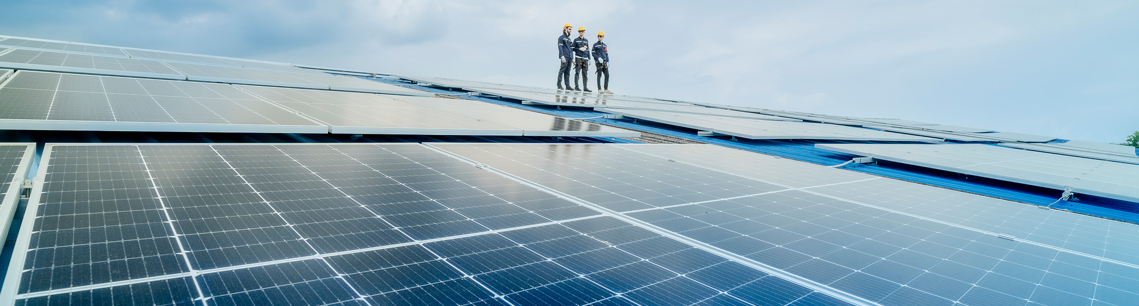 Großflächige Photovoltaikanlage unter blauem Himmel, mit drei Personen in Schutzkleidung, die auf den Solarpanels stehen und die Installation begutachten.