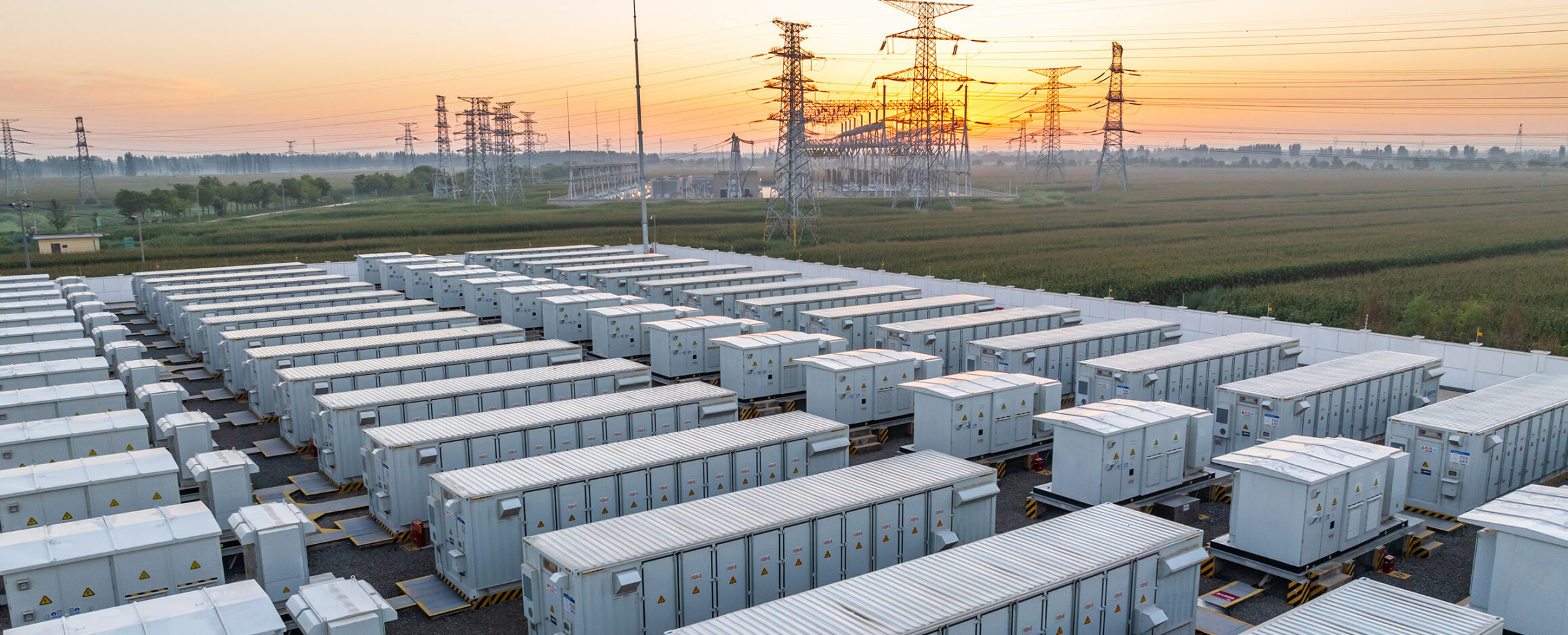 Große Batteriespeicheranlage mit zahlreichen Container-Einheiten, umgeben von Stromleitungen und Hochspannungsmasten bei Sonnenaufgang.