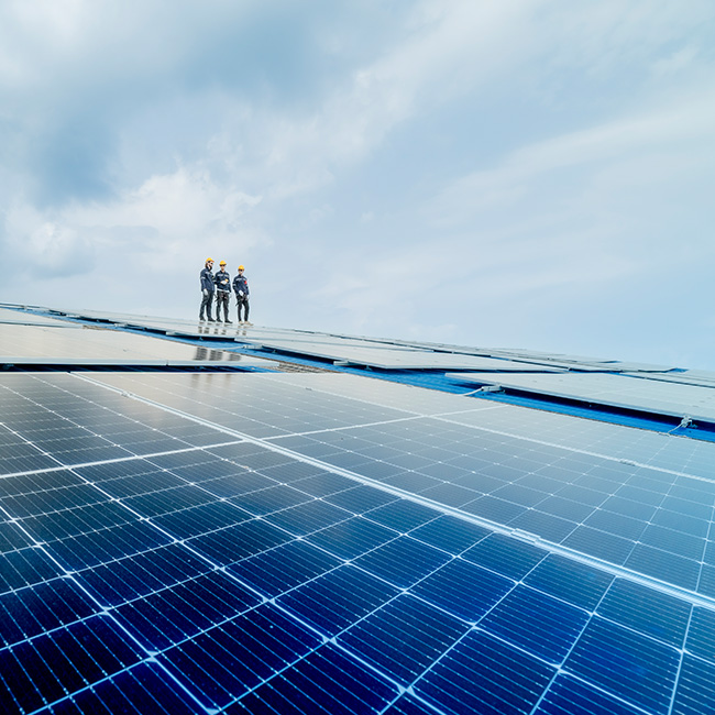 Großflächige Photovoltaikanlage unter blauem Himmel, mit drei Personen in Schutzkleidung, die auf den Solarpanels stehen und die Installation begutachten.