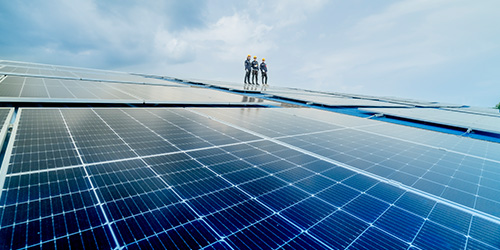Großflächige Photovoltaikanlage unter blauem Himmel, mit drei Personen in Schutzkleidung, die auf den Solarpanels stehen und die Installation begutachten.
